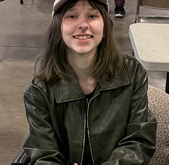 A photo of me sitting in a chair. I am wearing a dark green leather jacket and a light pink baseball cap. I am smiling at the camera